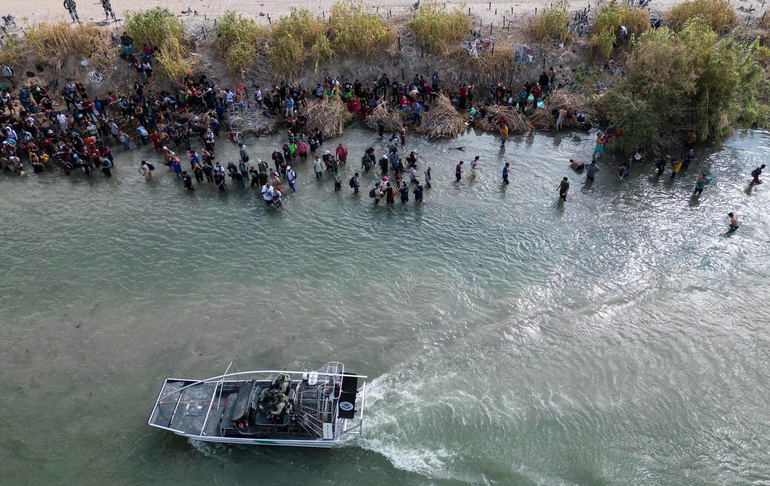 صورة جوية تظهر قارباً لحرس الحدود الأميركي يمر بمجموعة كبيرة من المهاجرين الذين ينتظرون عبور حاجز الأسلاك الشائكة إلى الولايات المتحدة في إيغل باس بولاية تكساس في 25 سبتمبر 2023 (أ.ف.ب)