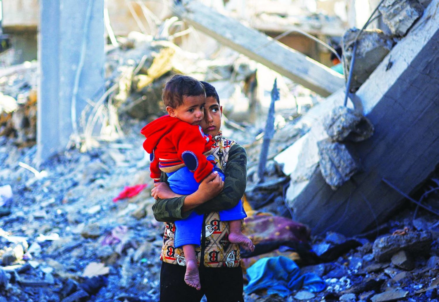 
صبي فلسطيني يحمل طفلاً وسط ركام منزل دمره قصف إسرائيلي في رفح جنوب قطاع غزة (رويترز)