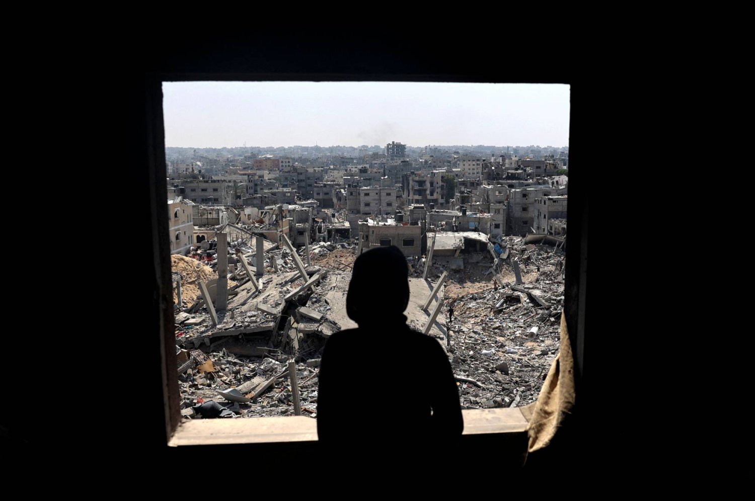 شاب فلسطيني ينظر عبر نافذة إلى مبانٍ دمرها القصف الإسرائيلي في خان يونس (أ.ف.ب)